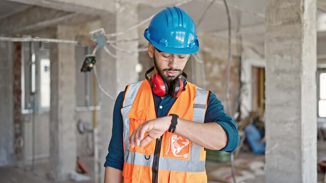 Young hispanic man builder looking at wrist watch at construction site
