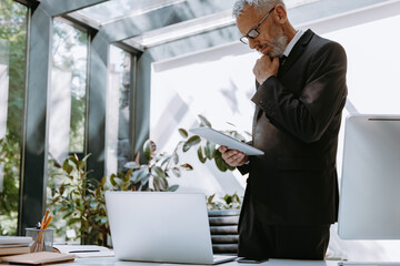 Concentrated mature businessman using digital tablet while standing near his working place in office
