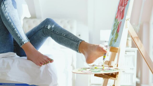 Focused female artist born without hands, squeezing the paint from the paint tube on the artist's palette with her legs at home. Actual life of an artist with Phocomelia syndrome 