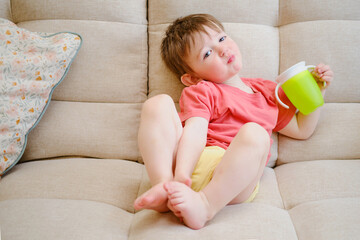 The non-spill cup is in the hands of the smiling child, who sits contentedly on the couch, drinking...