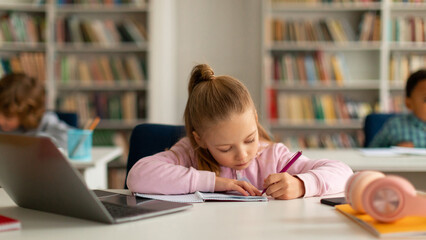 Focused caucasian pupil girl writing in copybook while using laptop computer, sitting at desk at elementary school