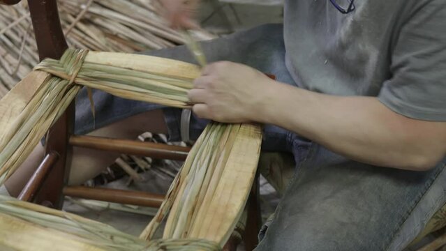 High angle of concentrated crop adult ethnic male in casual clothes looking down while sitting and weaving fiber rush chair seat in daylight in workshop