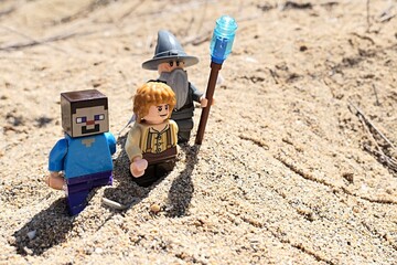 Fototapeta premium LEGO Minecraft figure of Steve exploring sandy beach with LEGO Lord Of The Rings wizard Gandalf and hobit Samwise Gamgee figures, summer daylight sunshine. Some arid foliage and beach in background. 