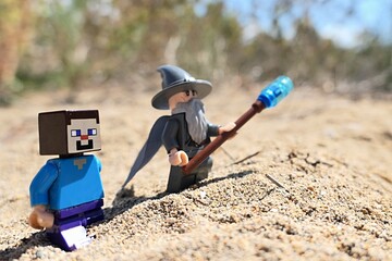 Fototapeta premium LEGO Minecraft figure of Steve walking on sandy beach with LEGO Lord Of The Rings wizard Gandalf figure, summer daylight sunshine. Some arid foliage and beach in background. 