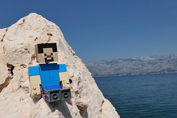 Obraz premium LEGO Minecraft large figure of main character Steve sitting on rocky cliff of Radanovac beach near Razanac, northern Dalmatia, Croatia. Velebit mountain in background, august bright sunshine. 