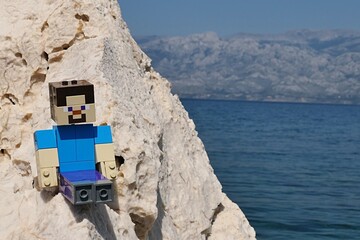 Fototapeta premium LEGO Minecraft large figure of main character Steve sitting on rocks of Radanovac beach near Razanac, northern Dalmatia, Croatia. Velebit mountain in background, august bright sunshine. 