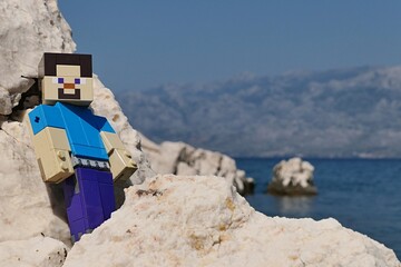 Obraz premium LEGO Minecraft large figure of main character Steve posing on rocks of Radanovac beach near Razanac, northern Dalmatia, Croatia. Velebit mountain in background, august bright sunshine. 