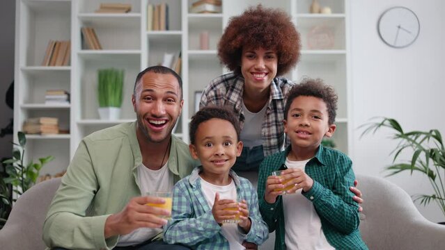 Portrait Of Joyful African American Spouses And Children Smiling At Camera While Drinking Fresh Juice In Home Interior. Young Pretty Family Woman Cuddling Kids And Excited Father From Behind