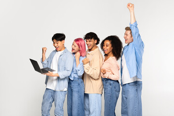 Five diverse friends holding laptop and shaking fists in joy, celebrating victory or reading great news online