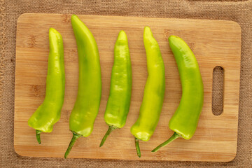 Several green chili peppers with wooden kitchen board on jute cloth, macro, top view.