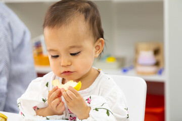 Toddler eating cut apple and sitting by the table in kindergarten