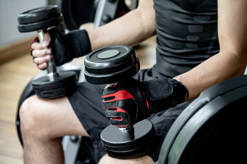 Athlete man in black sportswear wearing sport gloves sitting on workout bench at weightlifting station holding dumbbells prepare for lifting in fitness gym. Weight training and bodybuilding concepts