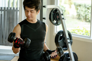 Asian sport man in black sportswear with sport gloves lifting dumbbells while sitting on bench at weightlifting station in fitness gym. Weight training and bodybuilding workout concepts