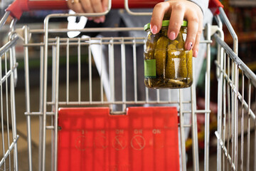 Close-up of a supermarket shopping cart and a woman's hand putting a jar of canned cucumbers into it.