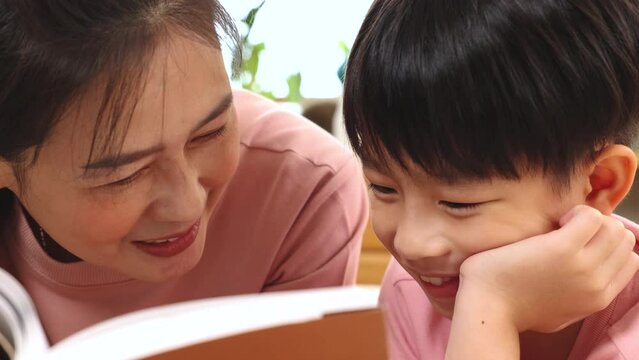 Asian beautiful mother reading fairy tale stories to adorable son lying down on bed, happy elementary school kid and single mom spend time together free time reading a book and tease in holiday.