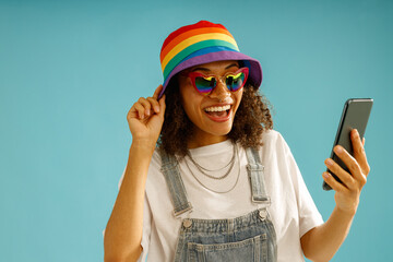 Happy woman in rainbow cap is holding mobile phone and looking at camera over blue studio background