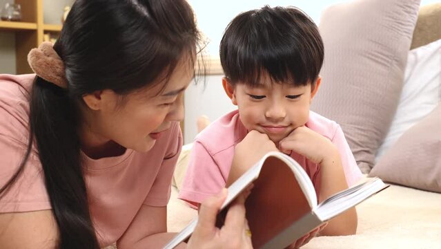 Asian beautiful mother telling fairy tale stories to handsome son laying on bed together, happy elementary school kid and single mom spend time free reading a book and tease in holiday.