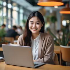 Fototapeta premium A beautiful and young Asian girl is sitting at a table with a laptop m smiling