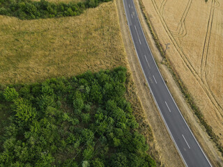 Aerial view of a rural asphalt road between ripe crop field and trees in the landscape 