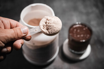 Male hand holds measuring spoon with whey protein powder above plastic jar on dark background