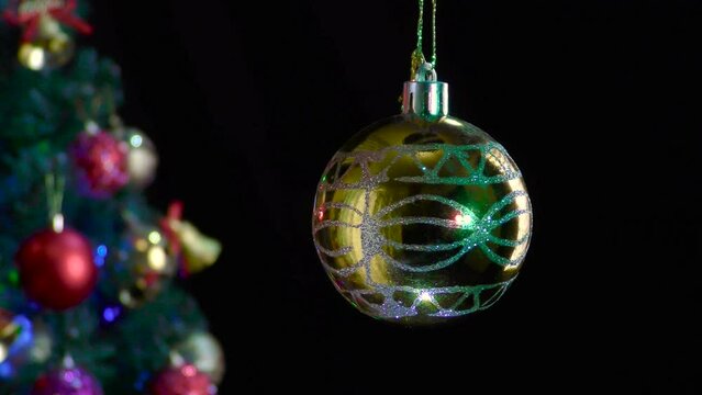 On a black background, a yellow glossy Christmas ball rotates near a decorated Christmas tree