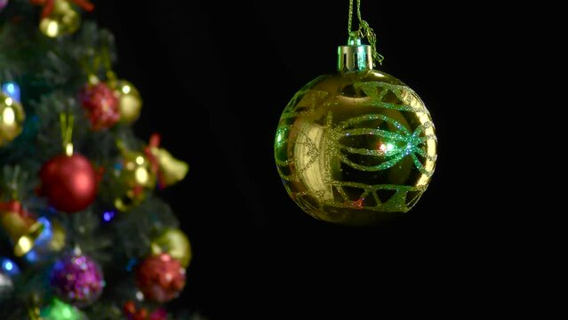 On a black background, a yellow Christmas ball with a pattern rotates near a decorated Christmas tree