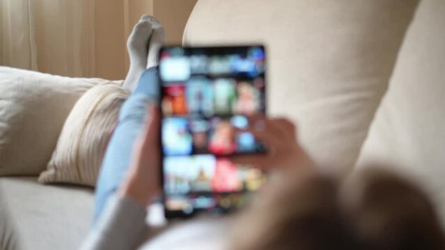 Unrecognizable person lying on a sofa in the living room looking for movies or television series to watch for pleasure. Woman choosing movies on internet streaming service. Blur