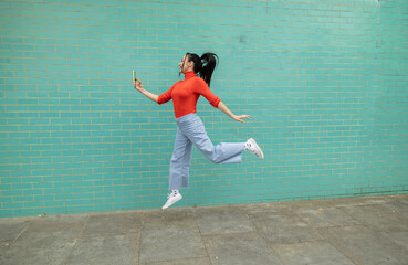 pretty woman jumping and making selfie light blue wall background