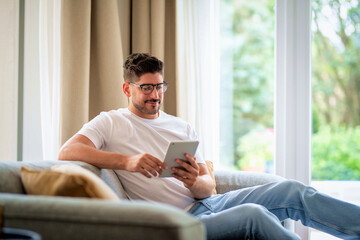 Middle-aged man sitting at home on the sofa and holding digital tablet in his hand - 643213944