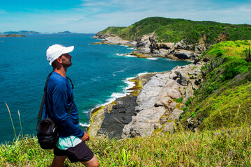 Man enjoying a view from above of the beautiful Praia das Conchas, near the city of Cabo Frio, with blue sea around, undergrowth, large rocks and mountains in the background - 80