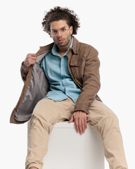 sexy man with curly hair and glasses adjusting beige jacket and sitting