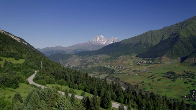 Panoramic aerial view of summer mountain valley landscape with green forest, snow peaks, small mountain village and sky (Ushba mountain, Svaneti region, Georgia)