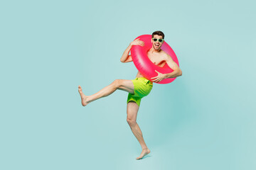 Full body young man wears green shorts swimsuit glasses relax near hotel pool hold inflatable ring lean back raise up leg isolated on plain blue background. Summer vacation sea rest sun tan concept.