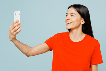 Young latin woman she wear orange red t-shirt casual clothes doing selfie shot on mobile cell phone post photo on social network isolated on plain pastel blue cyan background studio Lifestyle concept