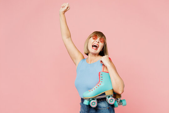 Overjoyed Elderly Blonde Woman 50s Years Old She Wearing Blue Undershirt Casual Clothes Do Winner Gesture Hold Roller Bladers Isolated On Plain Pastel Light Pink Background Studio. Lifestyle Concept.