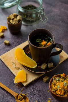 Ceramic cup with green tea with the addition of herbs, calendula flowers, candied mango and cane sugar on a plate, on a dark background.