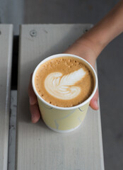 A kraft cup with cappuccino stands on a light bench, a hand takes it