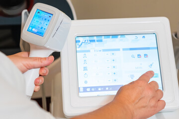Dermatologist stands holding device from laser epilation apparatus. Cosmetologist adjusted settings on device screen for laser epilation process