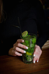 the hands of a young dark-haired girl in black clothes hold a non-alcoholic cool mojito on ice, a refreshing summer cocktail on a dark bar table
