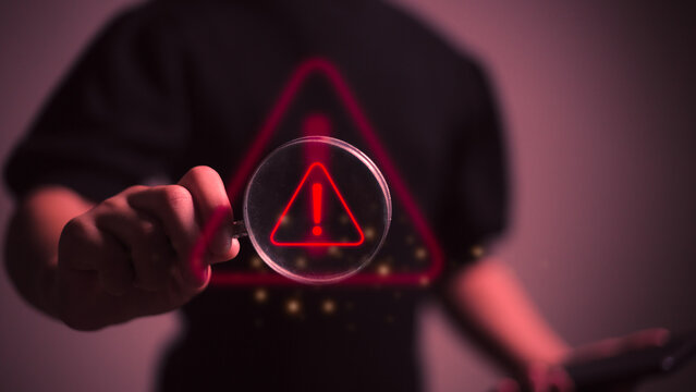 Hand holding magnifier glass with red triangle caution warning sign for focus notification error and maintenance concept.