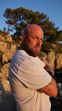 Striking portrait of a man with blue eyes and a beard, set against the picturesque landscape of Cap d'Antibes, France. His tattoos tell a story, embodying a blend of rugged charm and natural beauty