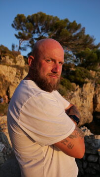 Striking portrait of a man with blue eyes and a beard, set against the picturesque landscape of Cap d'Antibes, France. His tattoos tell a story, embodying a blend of rugged charm and natural beauty