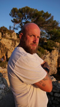 Striking portrait of a man with blue eyes and a beard, set against the picturesque landscape of Cap d'Antibes, France. His tattoos tell a story, embodying a blend of rugged charm and natural beauty