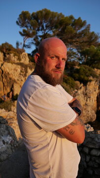 Striking portrait of a man with blue eyes and a beard, set against the picturesque landscape of Cap d'Antibes, France. His tattoos tell a story, embodying a blend of rugged charm and natural beauty