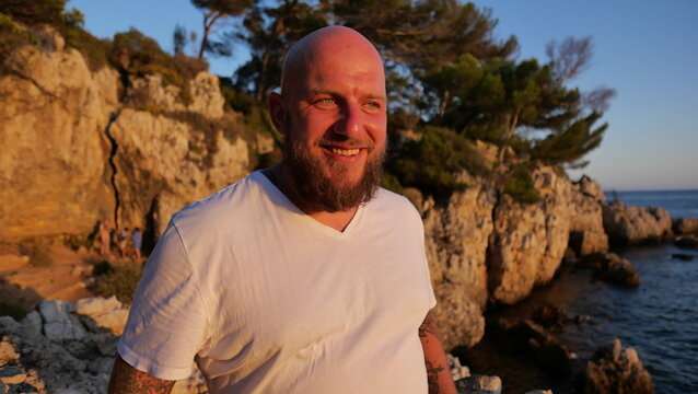 Striking portrait of a man with blue eyes and a beard, set against the picturesque landscape of Cap d'Antibes, France. His tattoos tell a story, embodying a blend of rugged charm and natural beauty