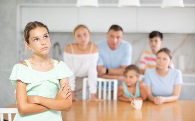 Offended pre-teen girl standing with arms crossed over chest at kitchen on background of her big family - obrazy, fototapety, plakaty