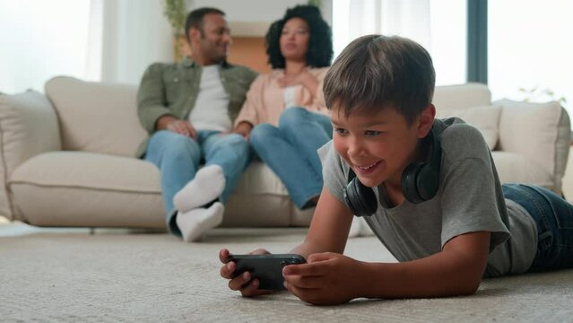 African American parents mother father talking on sofa little boy son child kid on floor play online video game on mobile phone internet addicted with smartphone happy multiracial family rest at home