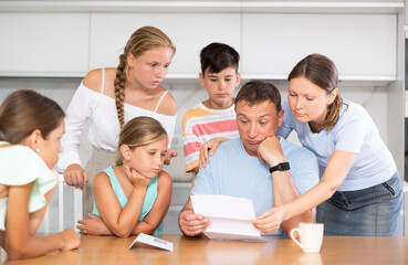 Fototapeta premium Large family is standing next to confused man sitting near kitchen table. Man reads letter and is upset by bad news from collector, unexpected test results, increase in loan rate
