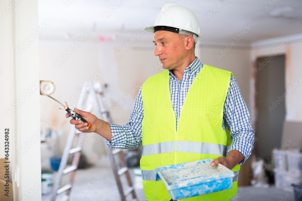 Wall mural Man builder with paint roller and tray painting wall in apartment.