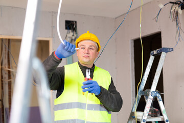 Repair man in yellow hardhat and vest working on electric wires during reconditioning works in apartment.